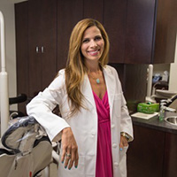 Sonia Massol-Burris smiles at the camera, wearing a magenta dress and a white coat, while she rests one arm on a dental table and the other hand is in the pocket of the coat.